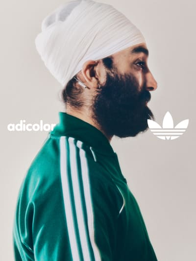A portrait of a man in a white turban and a green adicolor tracksuit while standing in front of a white backdrop. 