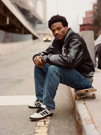 Tyshawn  looks to camera sitting on a skateboard, wearing a black leather jacket, jeans and tysawn II shoes