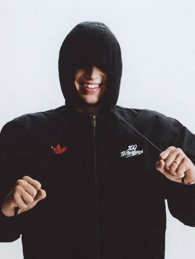 A man in a long sleeve shirt and cap from the adidas Originals x 100 Thieves collection strikes a pose in an empty white studio. 