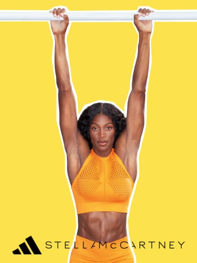 A woman with curly dark hair wearing an orange athletic top raises her arms triumphantly against a bright yellow background.