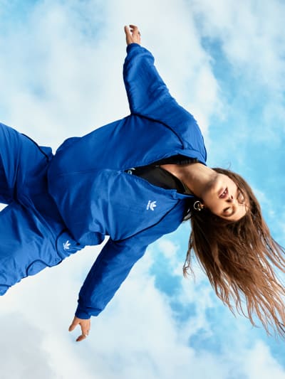 Female model wearing an adidas royal blue tracksuit with white adidas logo on the jacket and pants. Her long hair is flowing in the wind.
