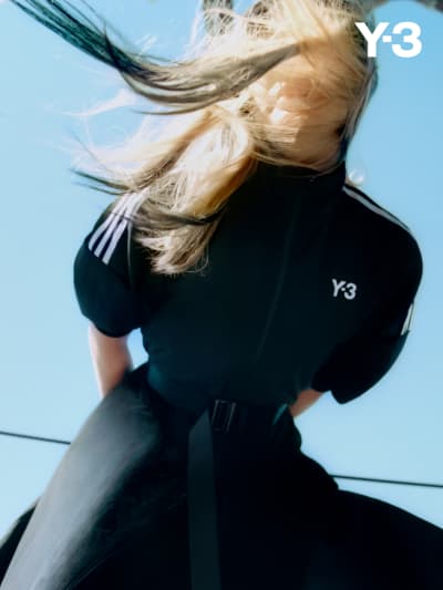 A woman wears a black Y-3 jacket in front of a blue sky while her blonde hair blows in the wind.