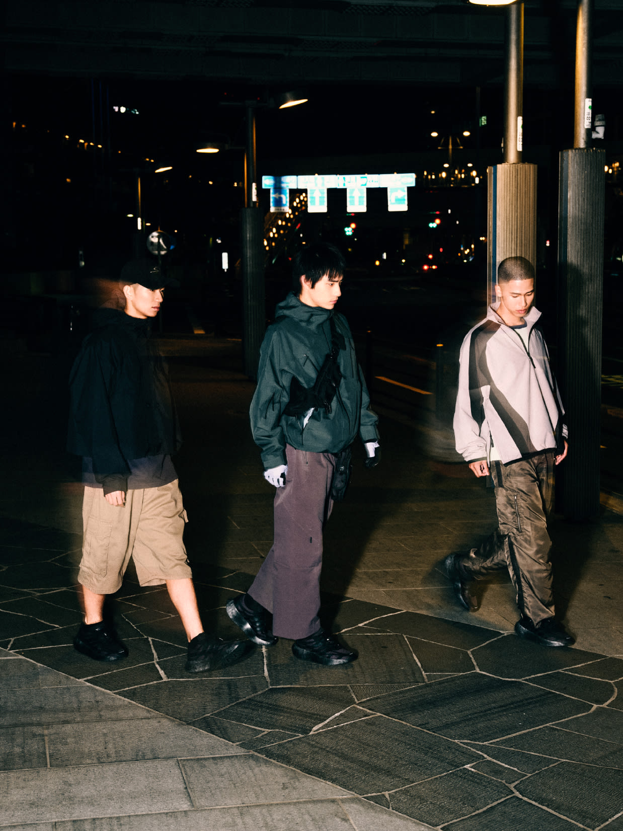 Three individuals, dressed in casual attire, stand on a paved surface in a dimly lit outdoor setting, with buildings and lights visible in the background.