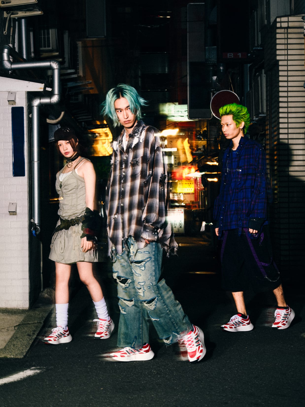 Three individuals with vibrant, dyed hair stand in a dimly lit urban setting, their casual attire and punk-inspired styles suggesting a youthful, alternative subculture.