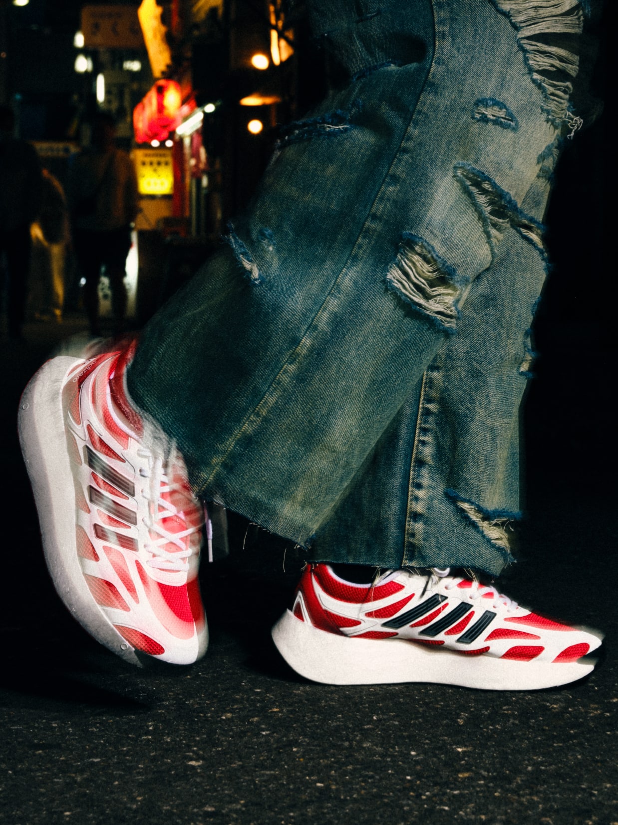 The image shows a pair of red and white sneakers on a person's feet, with the person wearing distressed denim jeans in the foreground, and a dimly lit urban setting visible in the background.