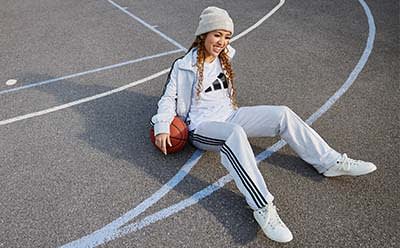 Relaxed, smiling woman is sitting on the floor of outdoor basketball court, leaning back onto a basketball for support.