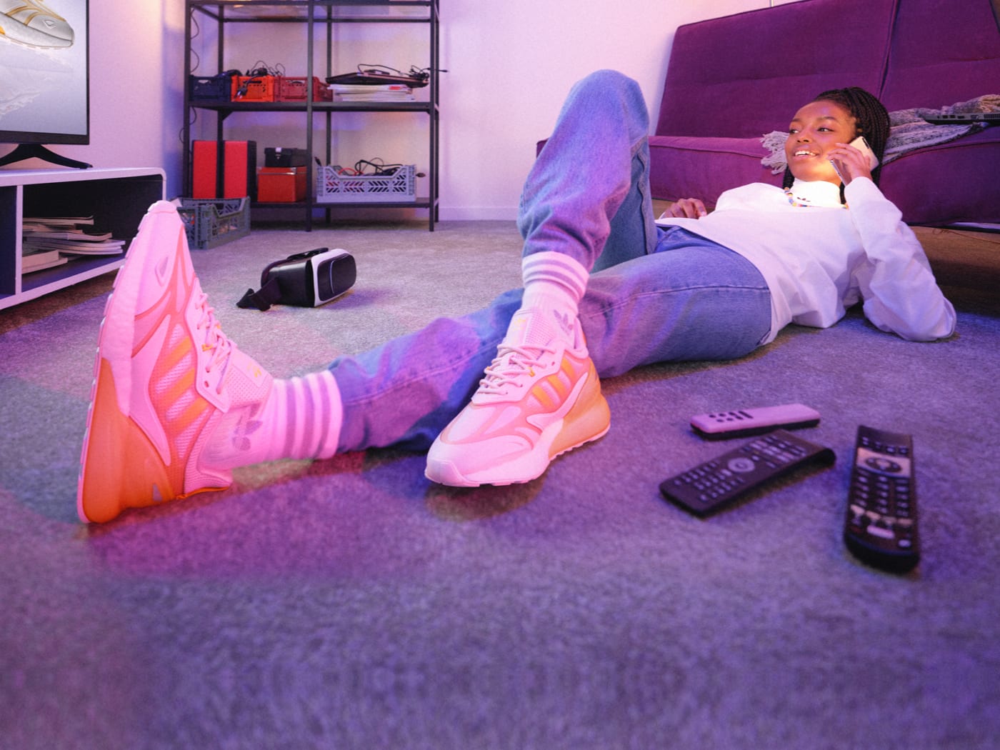 A model speaks on a cell phone that emits a glow against her cheek. She wears ZX 2K Boost 2.0 shoes that are also seen on a large TV screen.