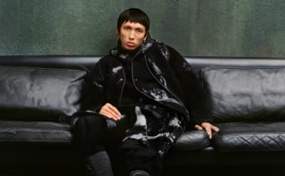 A model is pictured sitting on a leather sofa in a raw industrial building.