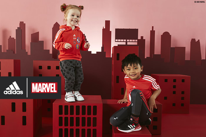 A child sat on a prop building while an infant is stood on another, both smiling wearing the adidas | MARVEL collection.