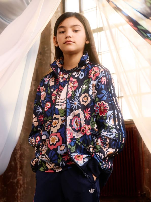 A young woman with long dark hair stands in a room with a sheer white curtain, wearing a colorful floral-patterned jacket.