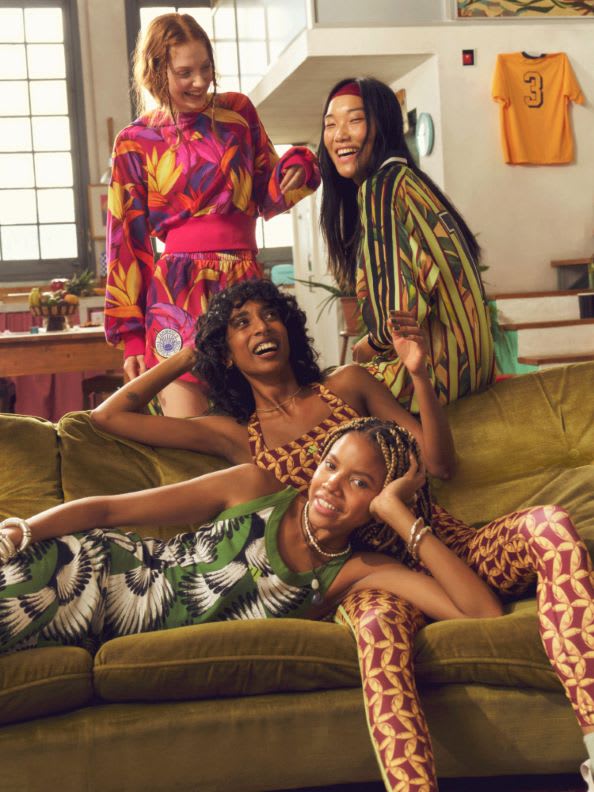 A group of young women, dressed in vibrant, patterned clothing, are relaxing and interacting on a couch in what appears to be a living room or home setting.