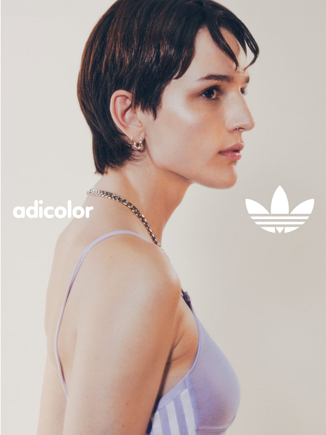 A portrait of a woman with short brown hair standing in front of a white wall while wearing a lavender adicolor sports bra and track pants.