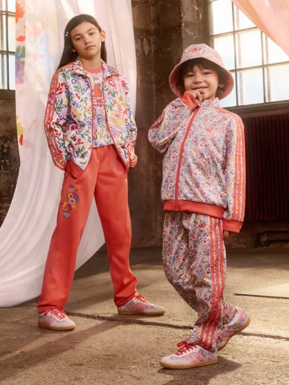 Two young girls wearing colorful, floral-patterned outfits stand in a rustic, indoor setting with a curtain-covered window in the background.