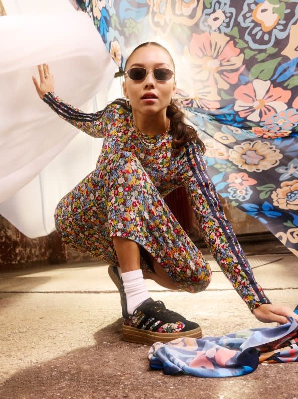 A young woman wearing a colorful floral print dress and sunglasses, posing on the floor in front of a vibrant floral-patterned backdrop.