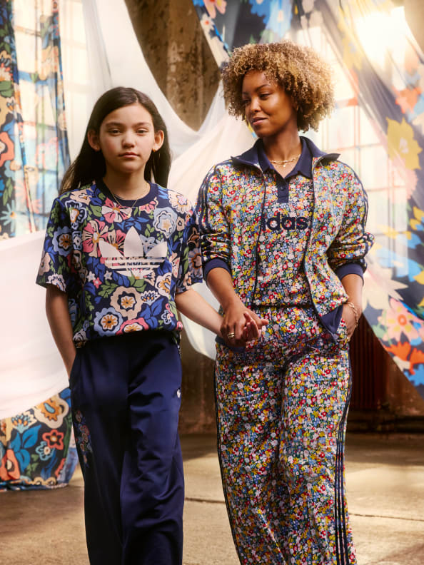 Two young women wearing colorful, patterned clothing stand together in front of a backdrop of vibrant, ornate textiles.