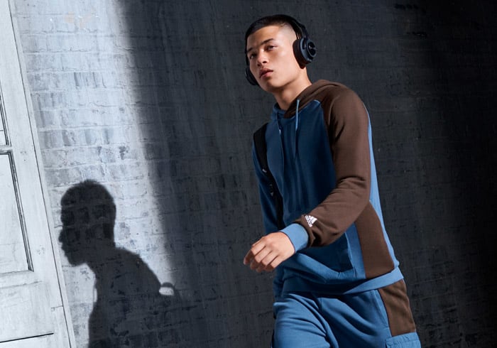 Image of male talent walking, wearing blue outfit with headphones on the head, against a grey wall, half shadow half light.