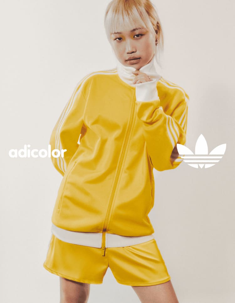 A woman with short brown hair sitting in front of a white backdrop while wearing a lavender adicolor sports bra and track pants.