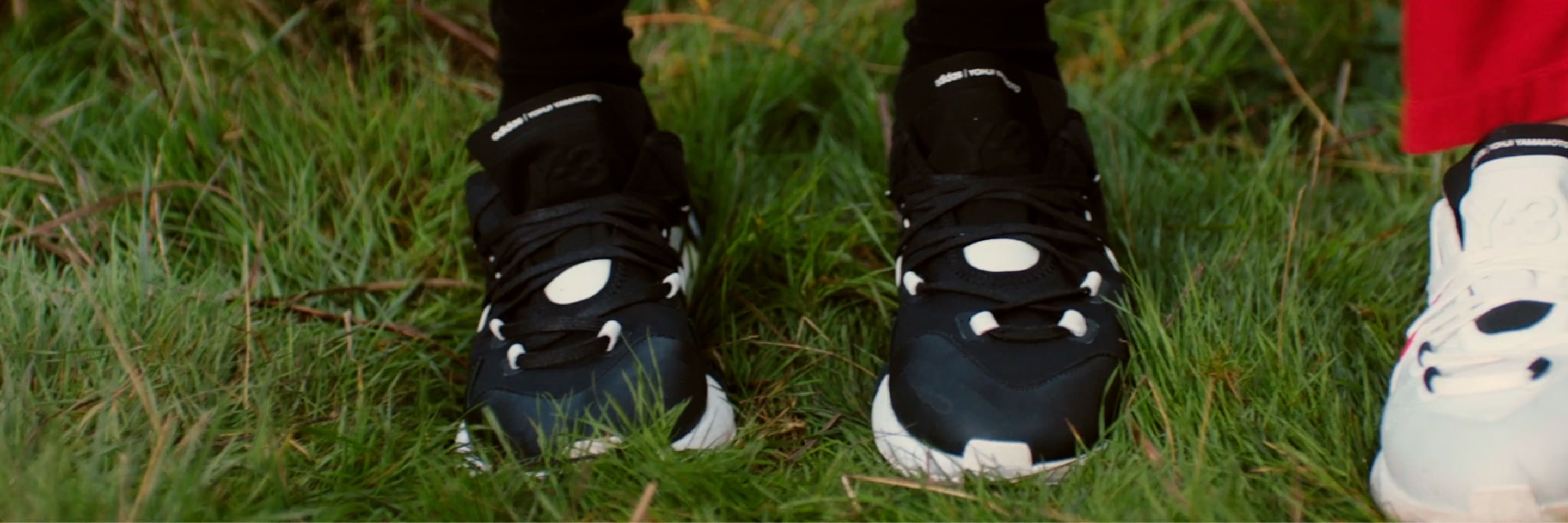 Video of two models walking in uncut grass, wearing the Y-3 IDOSO BOOST in both the black and white colorways.