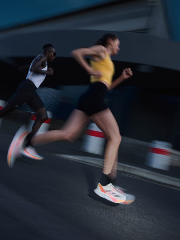 A man and woman are running on road, blurred in motion, wearing the Adizero Boston 12.