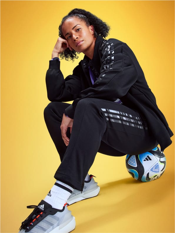 Soccer player Mary Fowler sits posed on a soccer ball, wearing a black adidas tracksuit and sneakers.