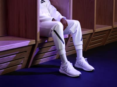 A man’s lower half wearing white adidas tracksuit top and matching bottoms and white trainers, sitting in a changing cubicle in a locker room.