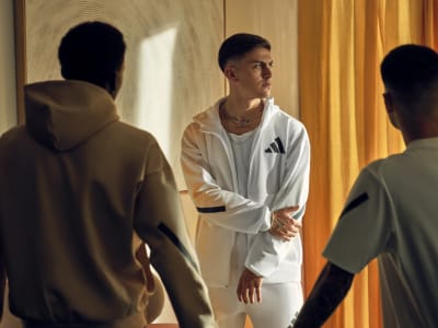 Footballer Paulo Dybala wears a white adidas tracksuit top and bottoms while standing with two other men.