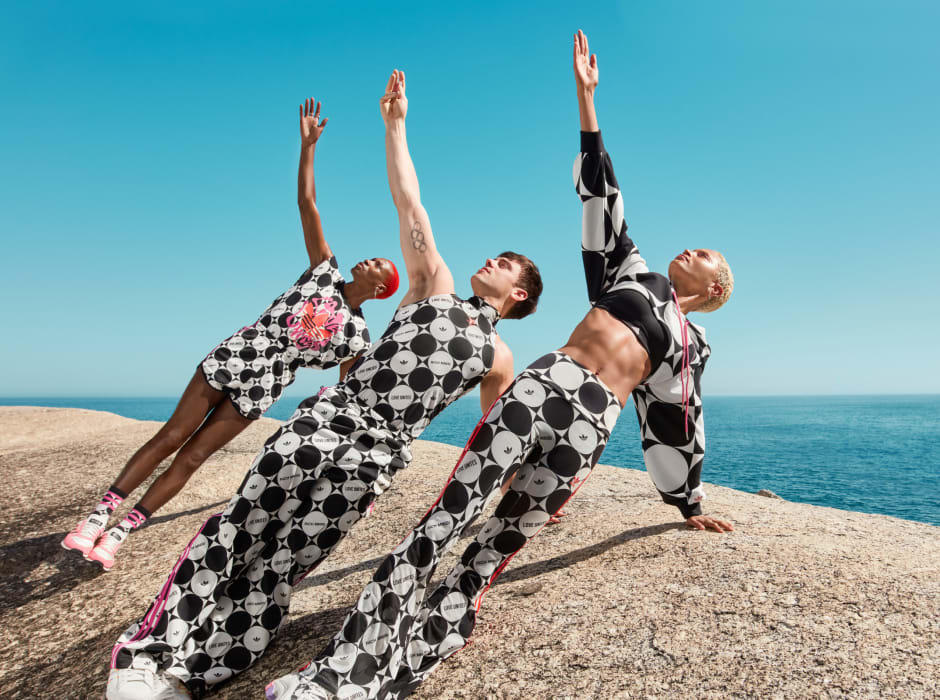 Tom Daley does a yoga pose, alongside two models, on a large stone overlooking an endless blue sky and ocean. They're all styled in apparel from the adidas x Rich Mnisi Pride Collection.
