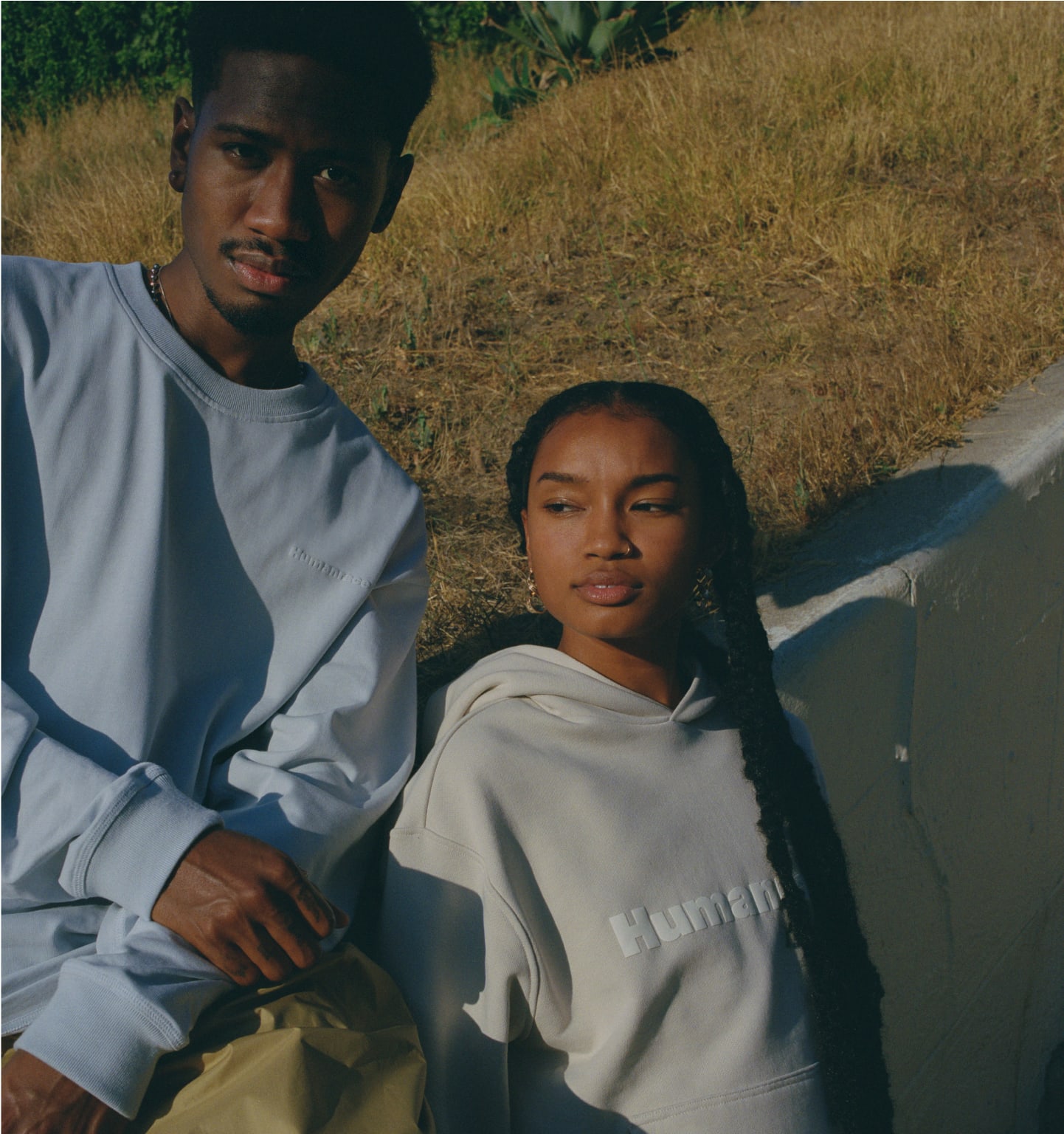 Two models wearing light blue Humanrace sweater and gray hoodie lean against wall