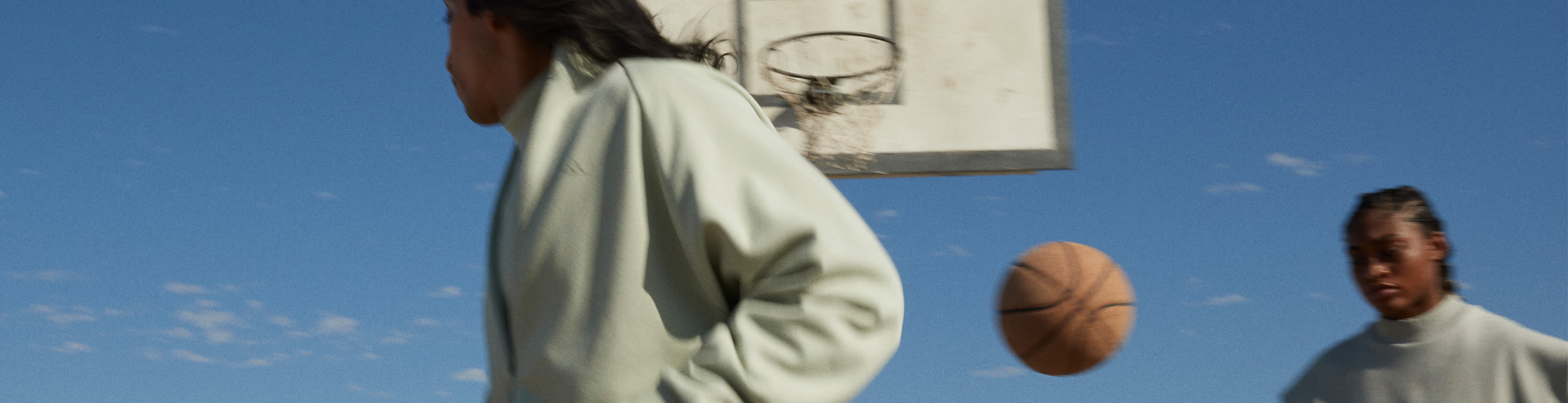 A waist-up view of two women with one women facing the back of the other women with a basketball in between them and blue sky and a basketball post in the background.