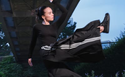 A woman in black Y-3 apparel stands in nature under a concrete bridge, kicking her feet high into the air. 