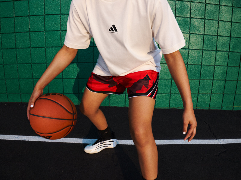 A person in a white t-shirt and red shorts is holding a basketball on a basketball court with a green tiled wall in the background.