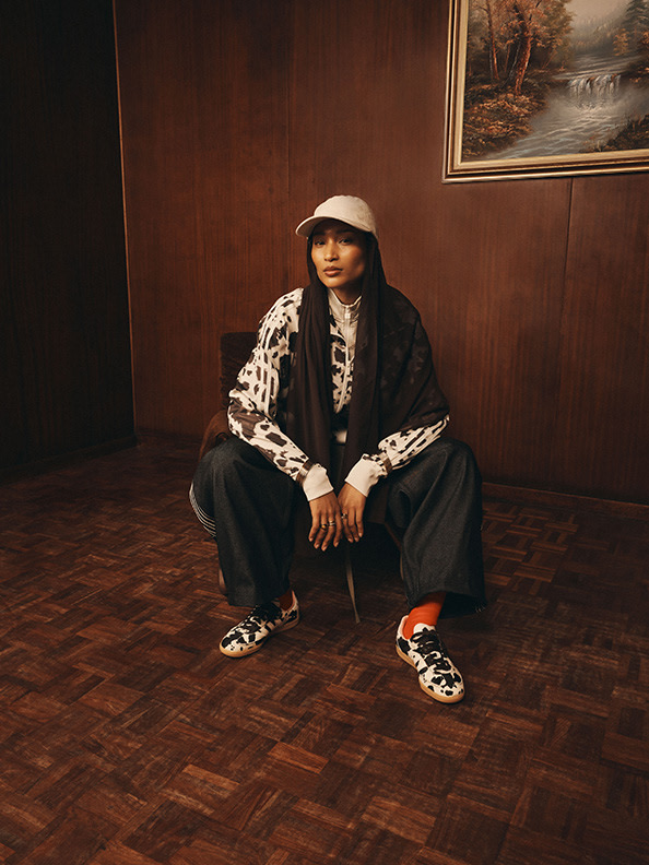 A person wearing a white hat, a patterned jacket, and red shoes sits on a wooden floor in front of a painting of a landscape on the wall.