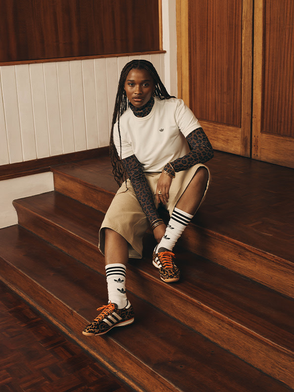 A person with dreadlocks sits on wooden steps, wearing a white top, beige shorts, and patterned socks with sandals, in a setting with wooden paneled walls.