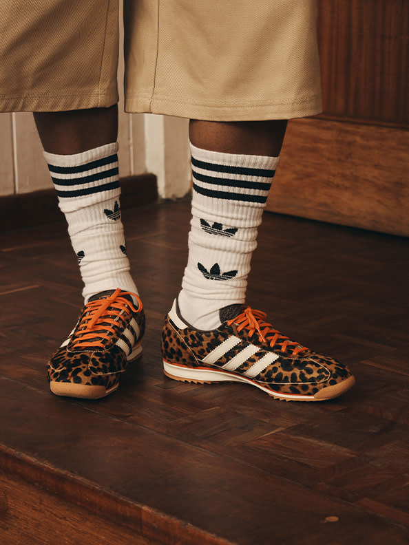 The image shows a pair of leopard-print sneakers with white socks featuring the Adidas logo, standing on a wooden floor.