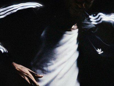 Person wearing a white tank top and a dark adidas Firebird jacket with the logo visible under dim lighting.