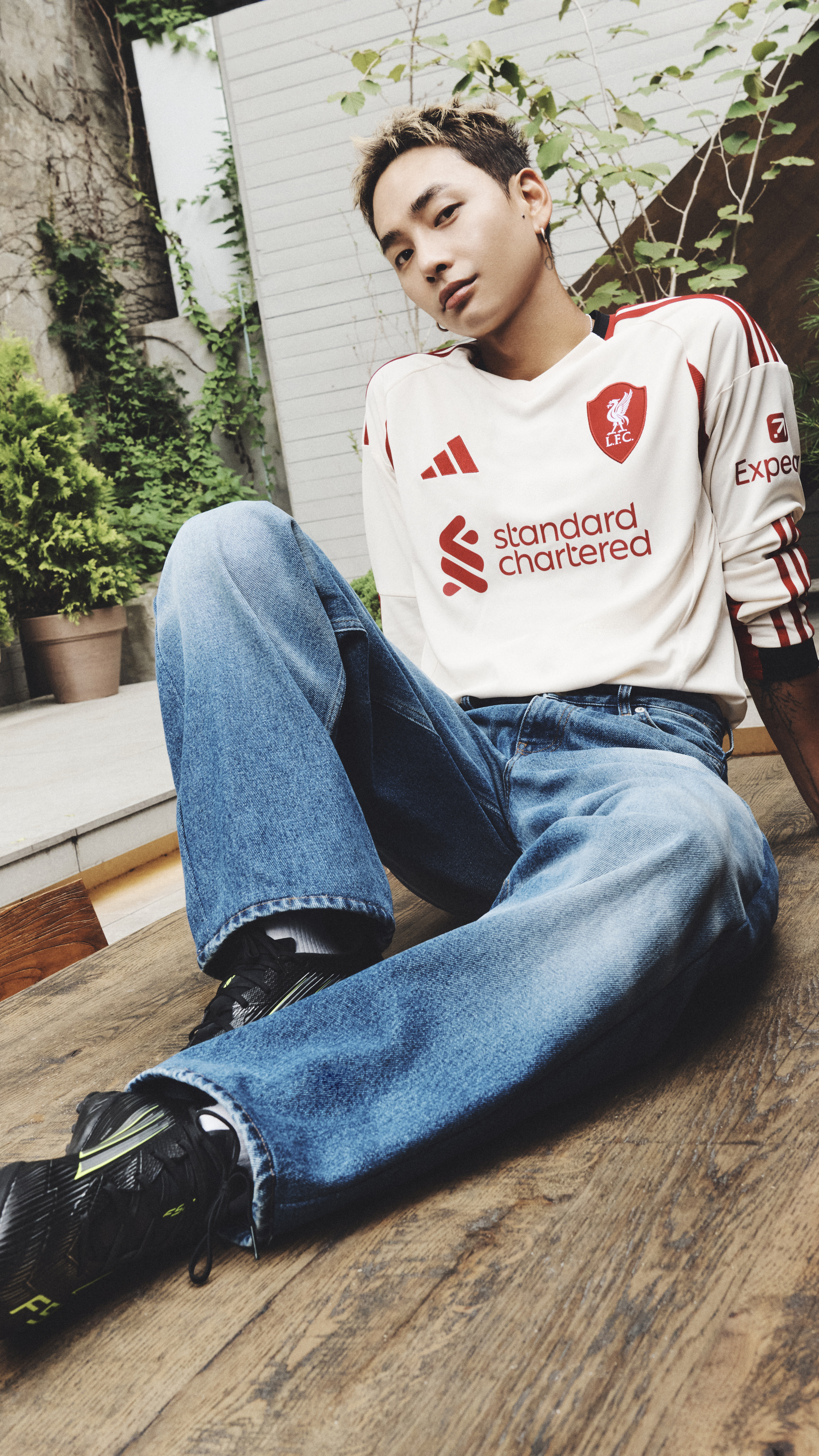 A young man wearing a Liverpool football club jersey is sitting on a wooden floor, surrounded by potted plants and a white wall in the background.