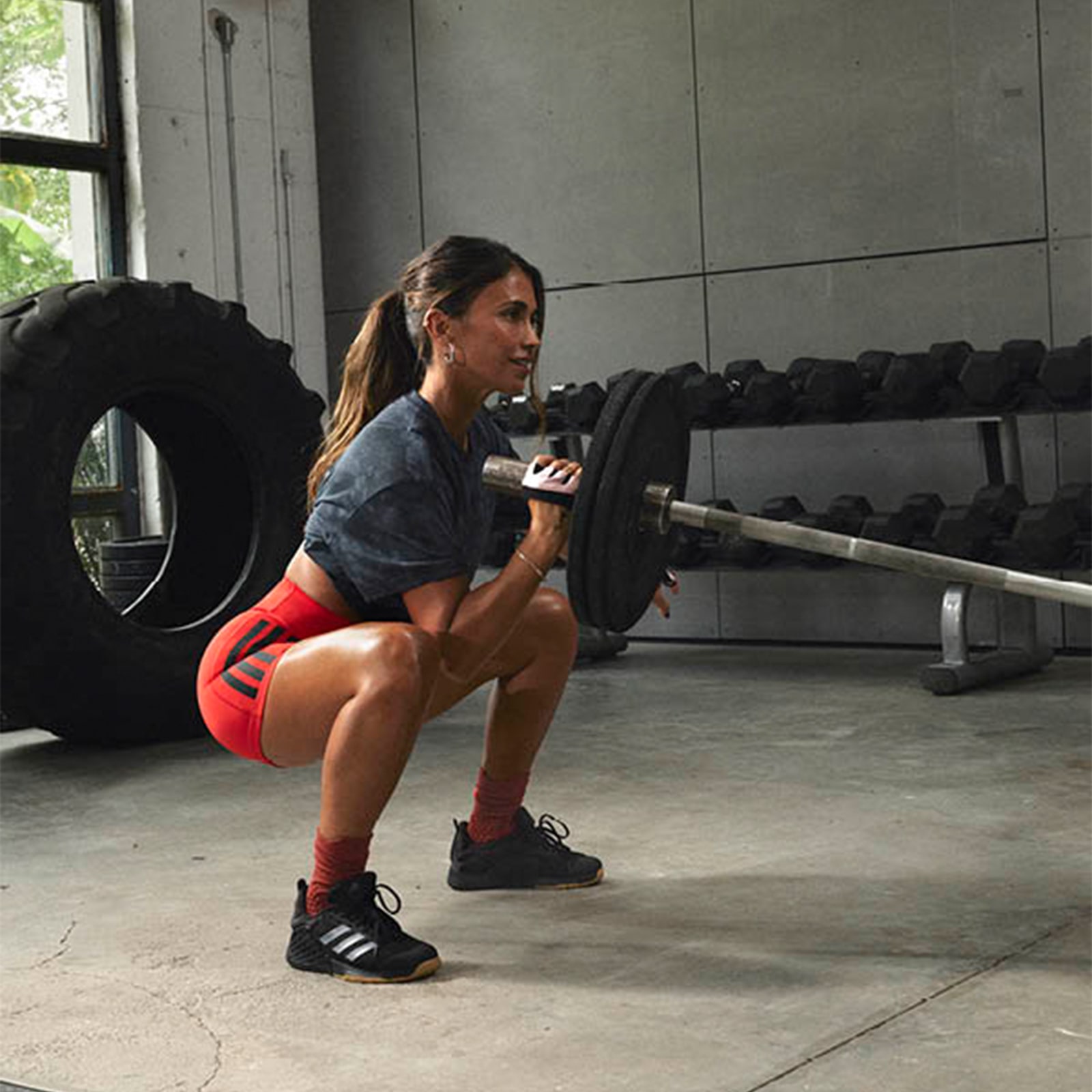 A woman in athletic clothing is performing a squat exercise with a barbell in a gym setting, surrounded by exercise equipment and a concrete floor.