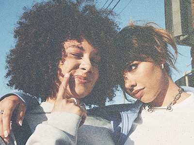 Deux jeunes femmes aux coiffures bien marquées, l'une à l'afro frisé, l'autre aux cheveux courts et sombres, sourient et posent ensemble dans un décor extérieur avec en arrière-plan bâtiments et lignes électriques.