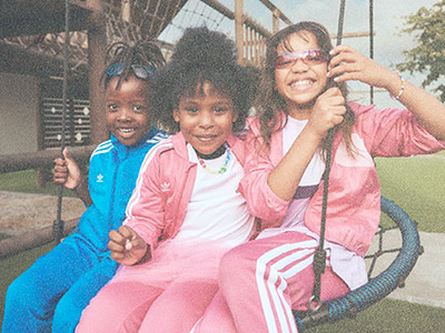 Trois jeunes filles, toutes habillées en bleu et en rose, sourient et posent ensemble sur une aire de jeux avec un cadre en bois en arrière-plan.