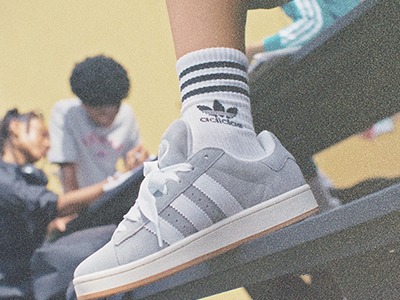 L'image montre les pieds d'une personne portant des sneakers adidas avec des chaussettes rayées, et les jambes d'une autre personne visibles en arrière-plan.