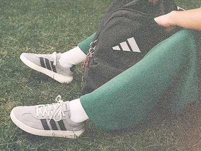 L'image montre les jambes et les pieds d'une personne portant des sneakers adidas sur un terrain en gazon.