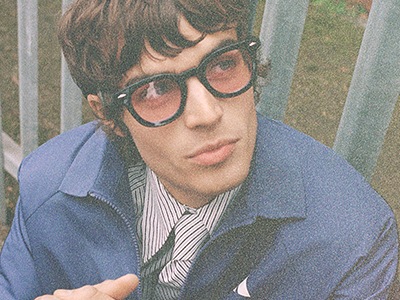 En ung man med mörkt hår och glasögon, iklädd en randig skjorta och en blå jacka, verkar vara utomhus mot en bakgrund av grönska.