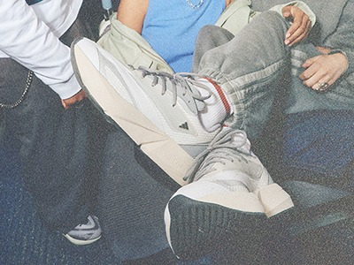 Bilden visar en närbild av en persons underkropp, med ett par grå sneakers och jeans synliga i förgrunden. Bakgrunden ser ut att vara en fullpackad miljö, med andras ben och fötter synliga runt ämnet.