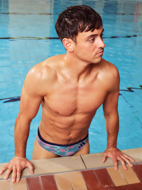 Un joven sin camiseta, con un cuerpo musculoso, está apoyado en el borde de una piscina, con un fondo de baldosas azules.