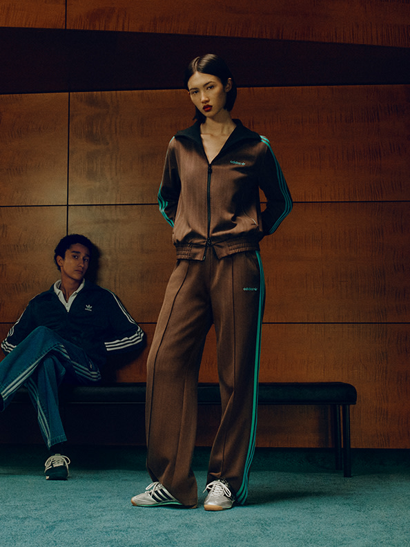 En person, der har en mørkfarvet jakke og bukser på, står i forgrunden, mens en anden person sidder på en bænk i baggrunden.