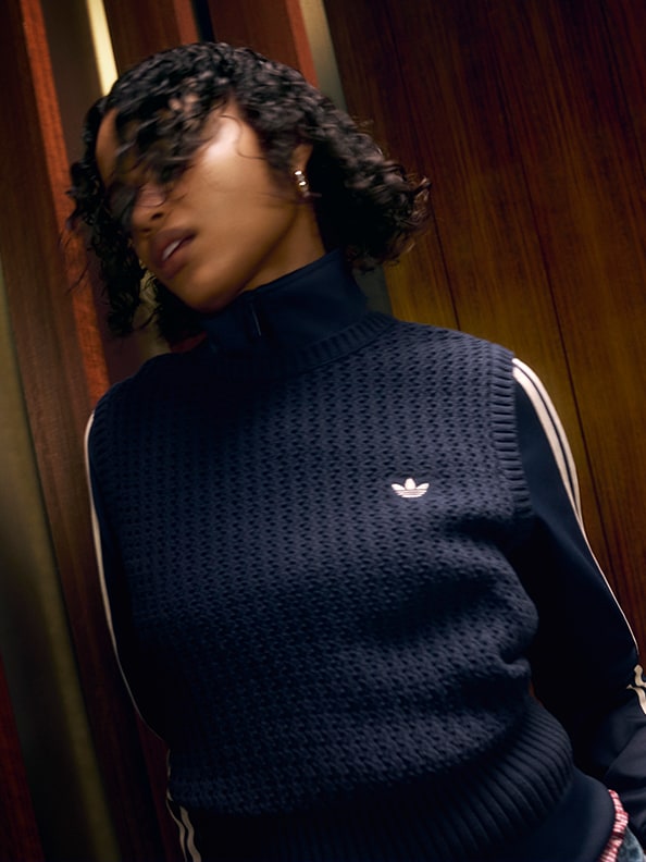 Billedet viser en person, der har en mørkeblå trøje på med adidas-logoet og står i et mørkt rum med trævægge.