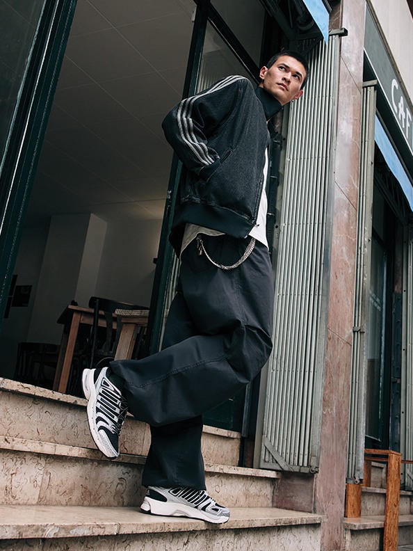 Une personne portant une tenue noire, y compris un sweat-shirt et un pantalon, se tient debout sur un escalier devant un bâtiment à la façade en verre.