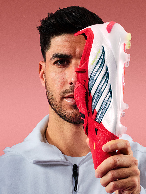 A young man with a serious expression holds a large, white and red athletic shoe in front of his face against a plain pink background.