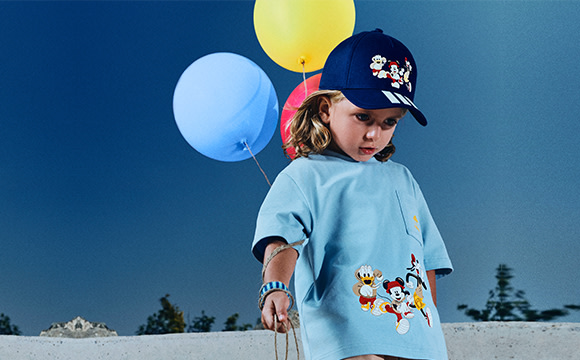A young person wearing a blue shirt and a baseball cap stands in a snowy outdoor setting, holding colorful balloons.