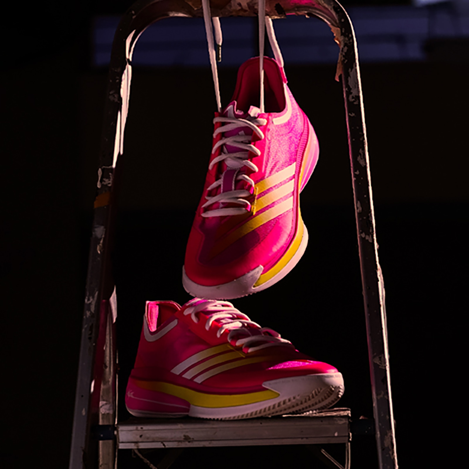 Vibrant pink and yellow athletic shoes are suspended on a metal rack against a dark background.